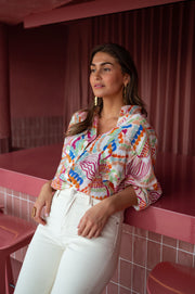 Chemise fluide manches trois-quarts portée par une femme, vue de trois-quarts, pantalon blanc, adossée à un comptoir