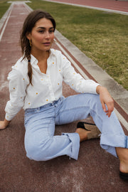 Chemise brodée à marguerites blanche portée par une femme, assise, piste rouge, jean clair