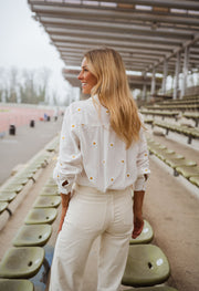 Chemise brodée à marguerites blanche portée par une femme, vue de dos, gradins, cheveux blonds