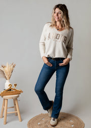 Pull brodé en maille fine porté par une femme, silhouette entière, jean, tabouret