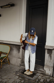 Gilet vichy sans manches porté par une femme, silhouette entière, casquette, pantalon blanc, mur blanc, de loin