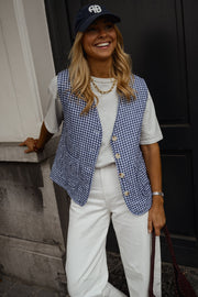 Gilet vichy sans manches porté par une femme, vue de trois-quarts, casquette bleu marine, pantalon blanc