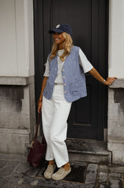 Gilet vichy sans manches porté par une femme, silhouette entière, casquette, pantalon blanc, porte noire