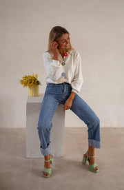 Gilet à boutons fleuris écru porté par une femme, assise, sur un cube blanc, jean