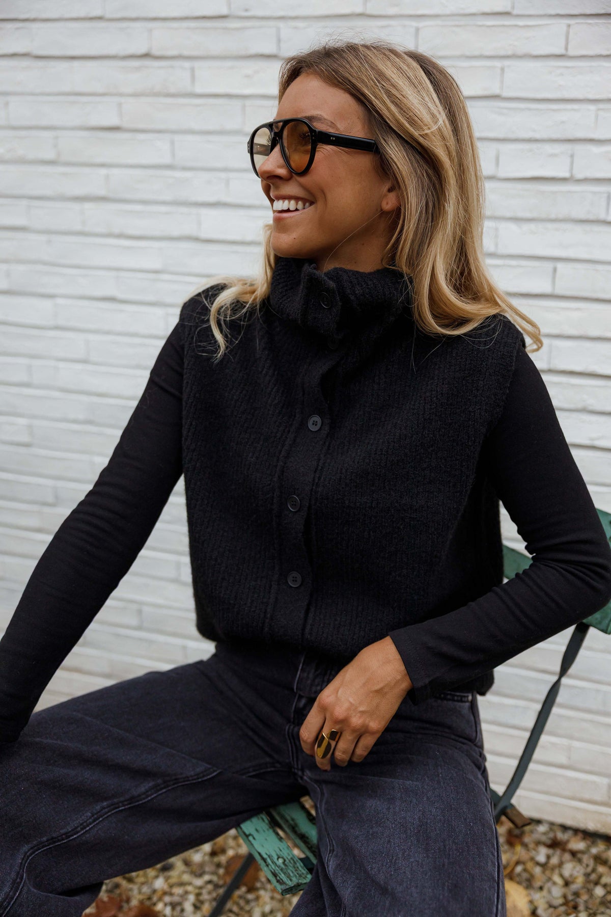 Gilet sans manches boutonné noir porté par une femme, assise, chaise verte, lunettes