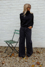 Gilet sans manches boutonné noir porté par une femme, vue de face, mur blanc, chaise verte