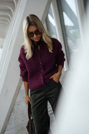 Gilet doux chaud à boutons bordeaux porté par une femme, vue de face, lunettes de soleil, piliers blancs
