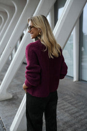 Gilet doux chaud à boutons bordeaux porté par une femme, vue de trois-quarts, piliers blancs, pantalon noir