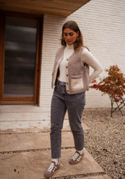 Gilet effet mouton et daim taupe porté par une femme, vue de face, mur blanc, jean gris