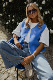Gilet à bordures fleuries bleu porté par une femme, assise, sur une chaise, lunettes de soleil