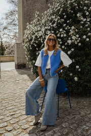 Gilet à bordures fleuries bleu porté par une femme, silhouette entière, jean, buisson fleuri, de loin