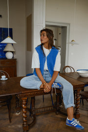 Gilet à bordures fleuries bleu porté par une femme, assise, à une table en bois, baskets bleues