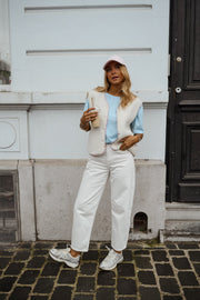 Gilet à bordures fleuries écru porté par une femme, silhouette entière, casquette, pantalon blanc, mur blanc, de loin