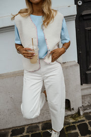 Gilet à bordures fleuries écru porté par une femme, vue de trois-quarts, gobelet à la main, pantalon blanc