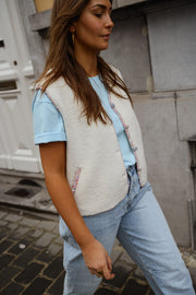 Gilet à bordures fleuries écru porté par une femme, vue de trois-quarts, jean clair, rue pavée