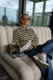 Gilet rayé à double zip porté par une femme, assise, sur un canapé, magazine à la main