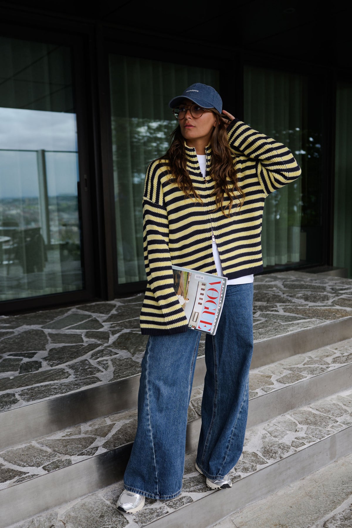 Gilet rayé à double zip porté par une femme, silhouette entière, casquette, sur un sol en pierre