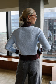 Gilet torsadé à col montant porté par une femme, vue de dos, près d'une fenêtre