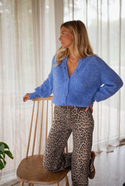 Gilet doux boutonné bleu porté par une femme, vue de face, chaise en bois, pantalon léopard