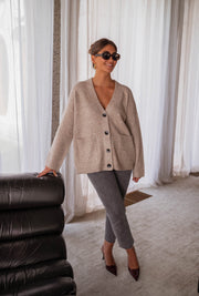 Gilet long droit à poches beige porté par une femme, silhouette entière, canapé noir, escarpins