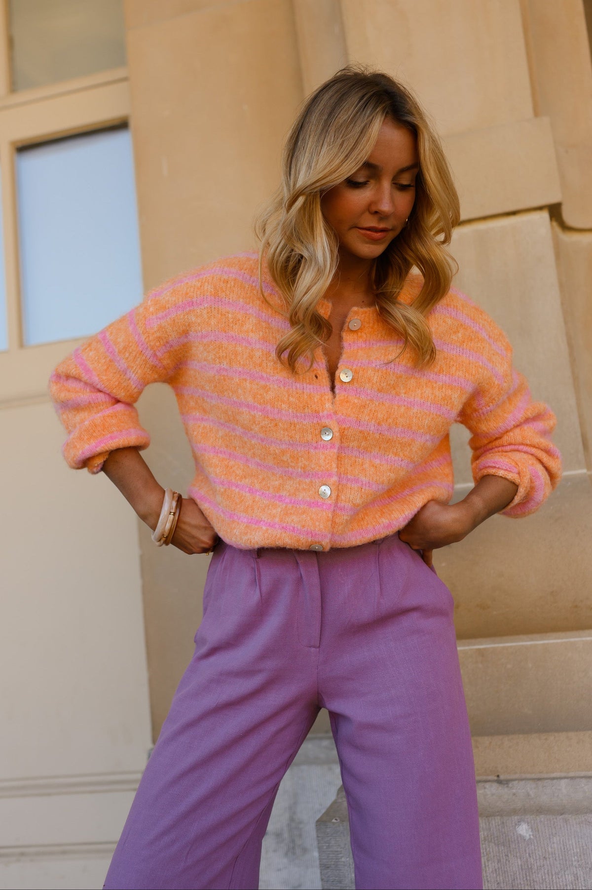 Gilet doux légèrement ample porté par une femme, vue de trois-quarts, pantalon violet, mains sur les hanches