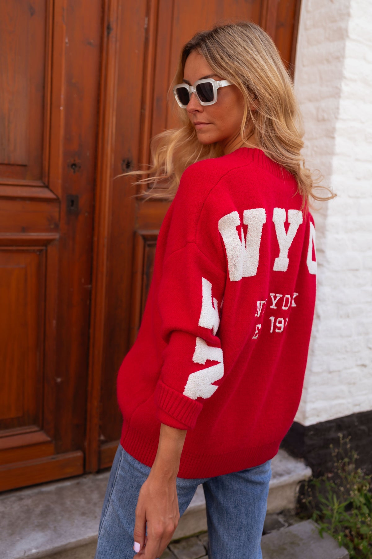 Gilet ample boutonné rouge porté par une femme, vue de dos, lettres blanches, porte en bois