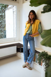 Gilet court à poches jaune porté par une femme, silhouette entière, près d'une fenêtre, plantes