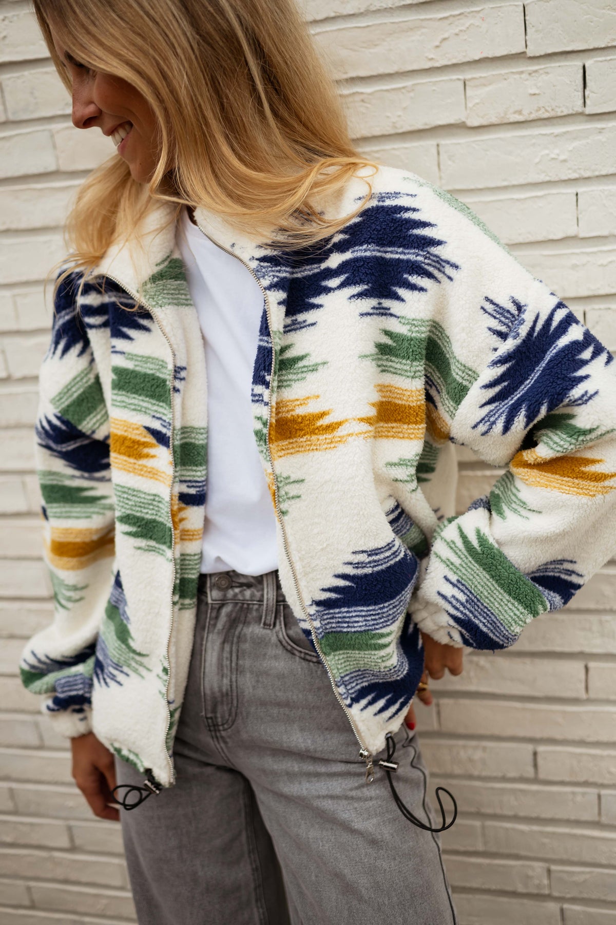 Gilet polaire zippé à motifs écru porté par une femme, vue de face, mur blanc, jean gris