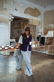 Gilet mouton court sans manches mauve porté par une femme, vue de face, comptoir du café, casquette