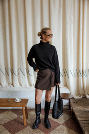 Jupe-short à carreaux porté par une femme, silhouette entière, bottes noires, sac cabas, fond rideau