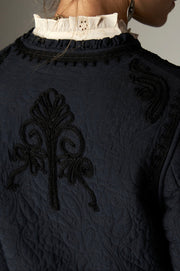 Gros plan sur le motif décoratif et la bordure de l’encolure au dos de la veste sombre.