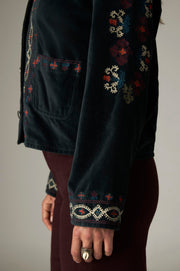 Détail des broderies multicolores sur la manche, le poignet et la poche de la veste sombre.