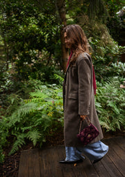 Manteau long doublé à épaulettes taupe porté par une femme, vue de trois-quarts, feuillage, sac bordeaux