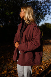 Manteau ample imperméable bordeaux porté par une femme, vue de trois-quarts, sous-bois, lunettes