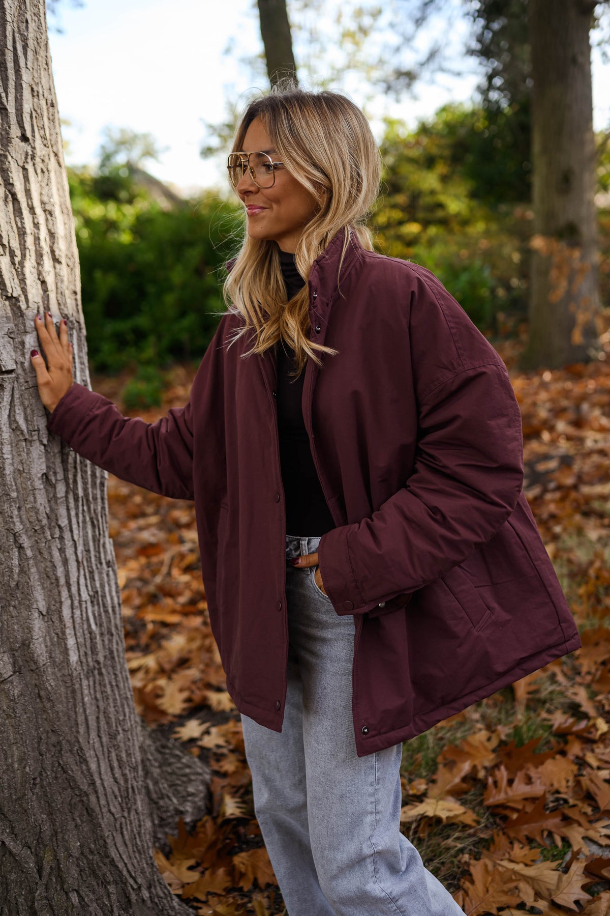 Manteau ample imperméable bordeaux porté par une femme, vue de trois-quarts, tronc d'arbre, jean gris