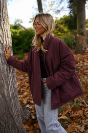 Manteau ample imperméable bordeaux porté par une femme, vue de trois-quarts, tronc d'arbre, jean gris