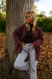 Manteau ample imperméable bordeaux porté par une femme, vue de trois-quarts, tronc d'arbre, jambe pliée