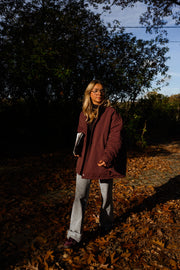 Manteau ample imperméable bordeaux porté par une femme, vue de profil, feuilles d'automne, jean gris