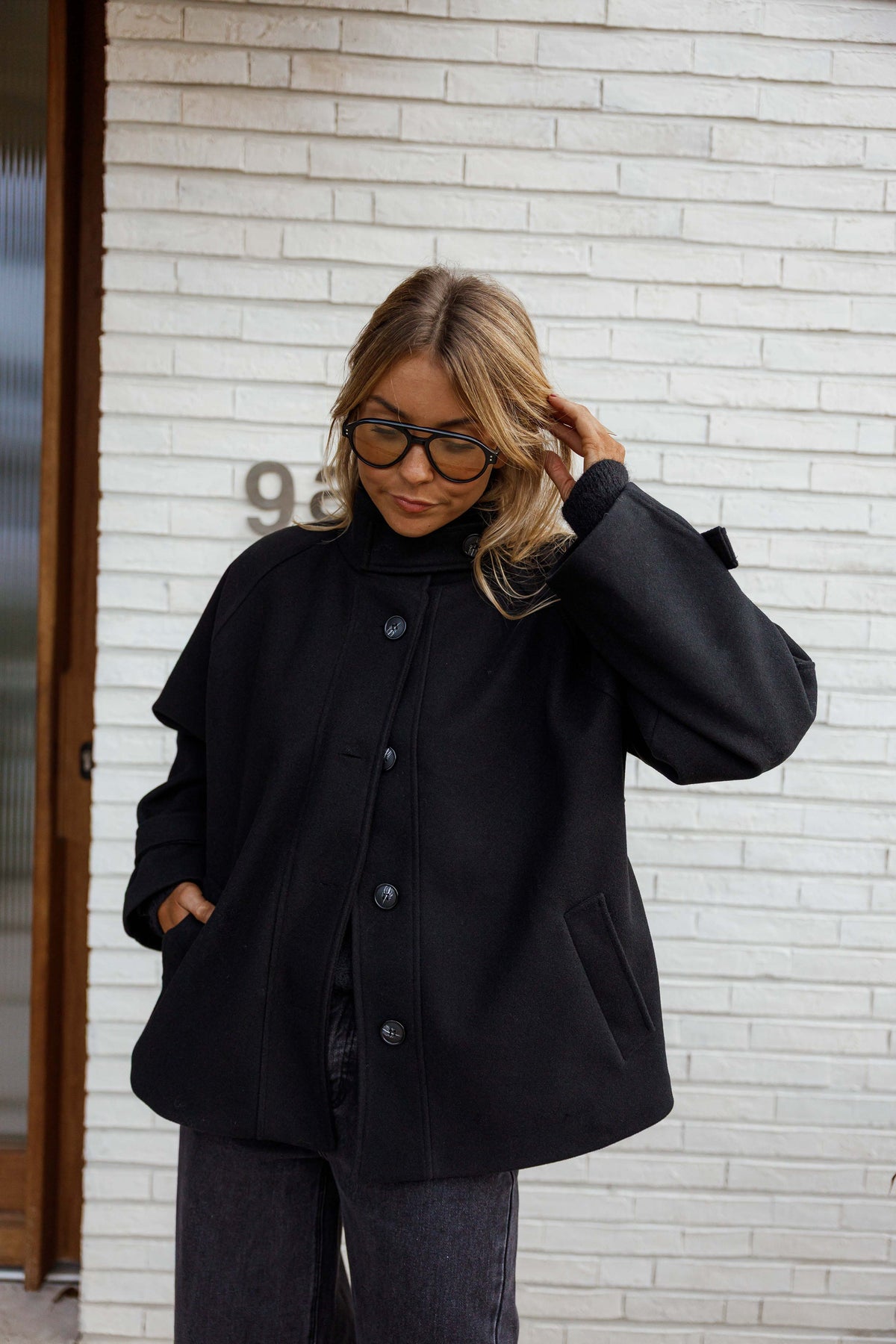 Manteau à col montant boutonné noir porté par une femme, vue de trois-quarts, porte en bois, lunettes