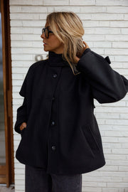 Manteau à col montant boutonné noir porté par une femme, vue de trois-quarts, mur blanc, bras en arrière