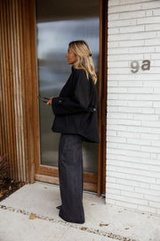 Manteau à col montant boutonné noir porté par une femme, vue de dos, porte vitrée, pantalon gris