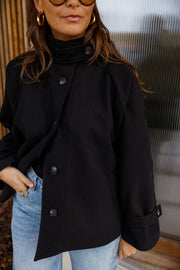 Manteau à col montant boutonné noir porté par une femme, vue de face, porte en bois, jean bleu