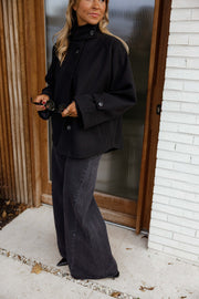 Manteau à col montant boutonné noir porté par une femme, vue de trois-quarts, porte en bois, téléphone