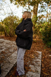 Manteau à capuche imperméable noir porté par une femme, vue de profil, allée pavée, bras croisés