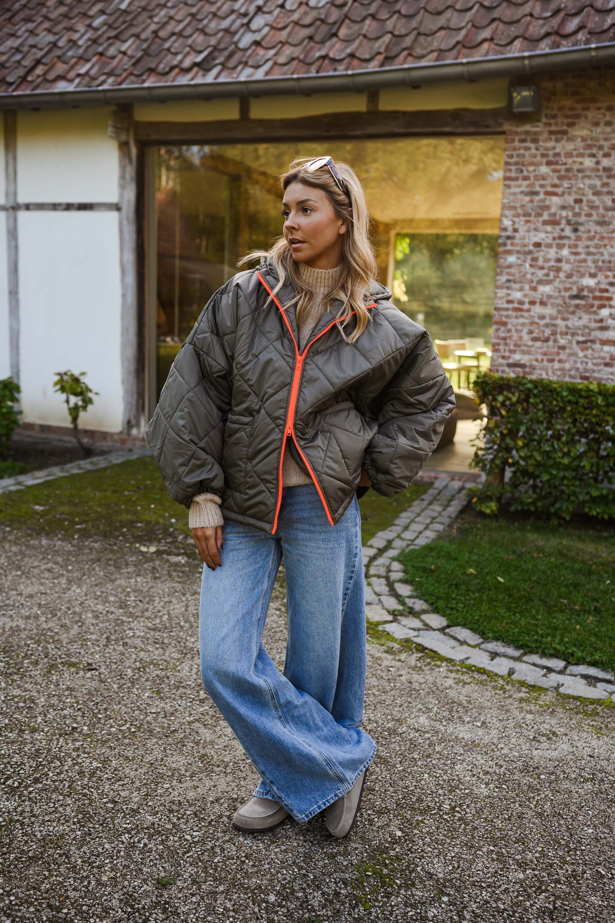 Manteau imperméable matelassé kaki porté par une femme, vue de face, mur en briques, fermeture orange