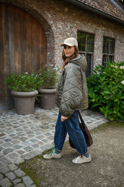 Manteau imperméable matelassé kaki porté par une femme, vue de trois-quarts, pots de fleurs, casquette beige