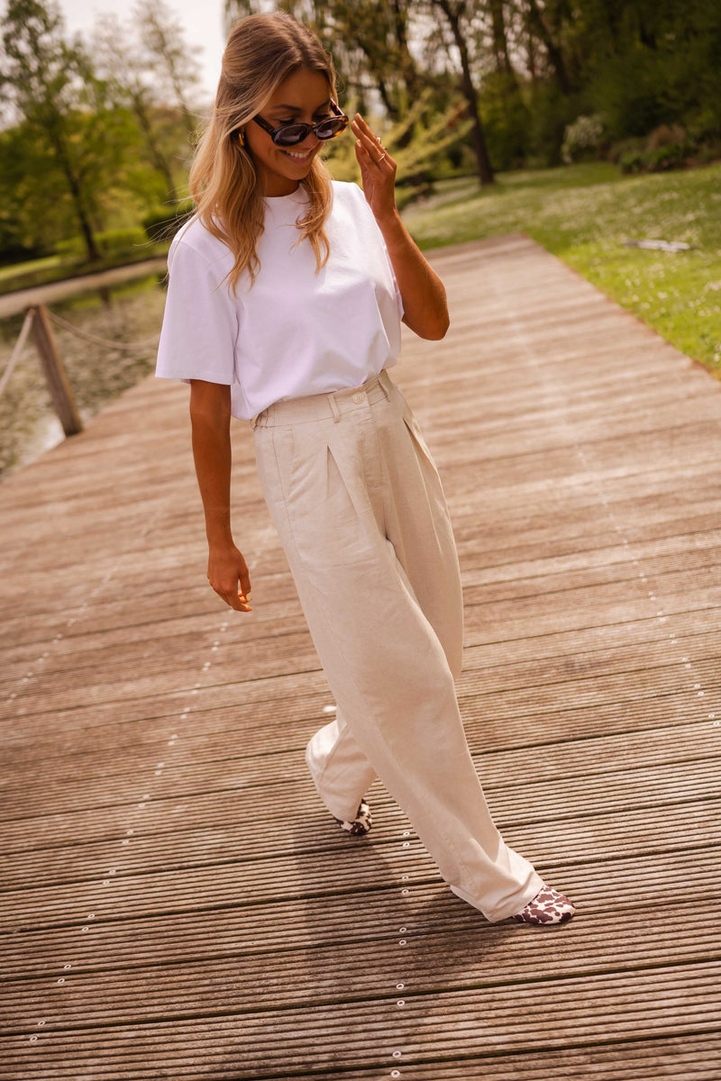 Pantalon fluide en lin beige porté par une femme, silhouette entière, ponton en bois, pantalon beige