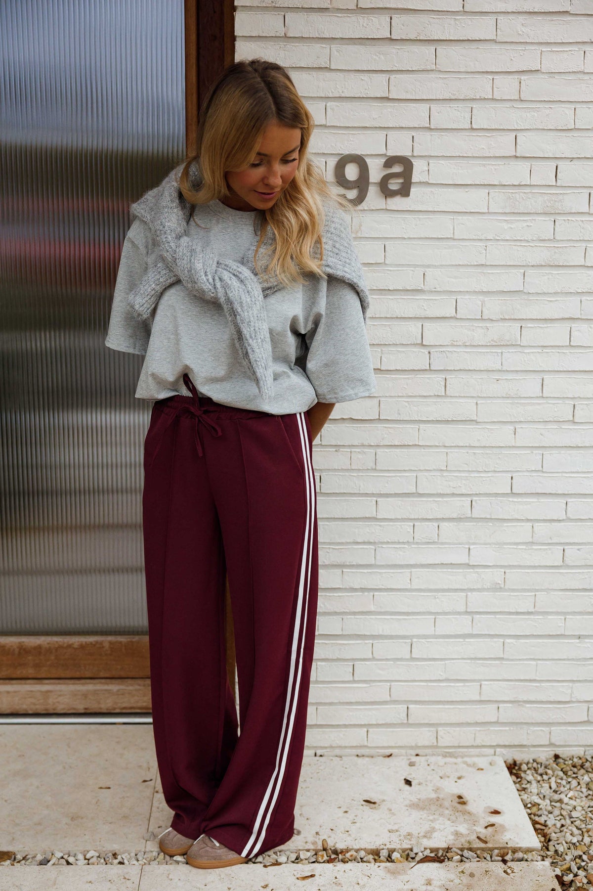 Pantalon à bandes latérales porté par une femme, silhouette entière, pull noué sur les épaules, mur blanc