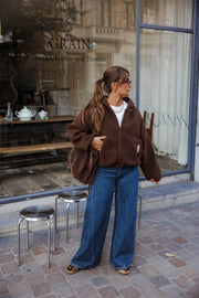 Veste polaire zippée à capuche marron portée par une femme, en pied, jean large bleu, tabourets métalliques, trottoir pavé