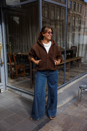 Veste polaire zippée à capuche marron portée par une femme, vue de face, mains dans les poches, collier, vitrine de café